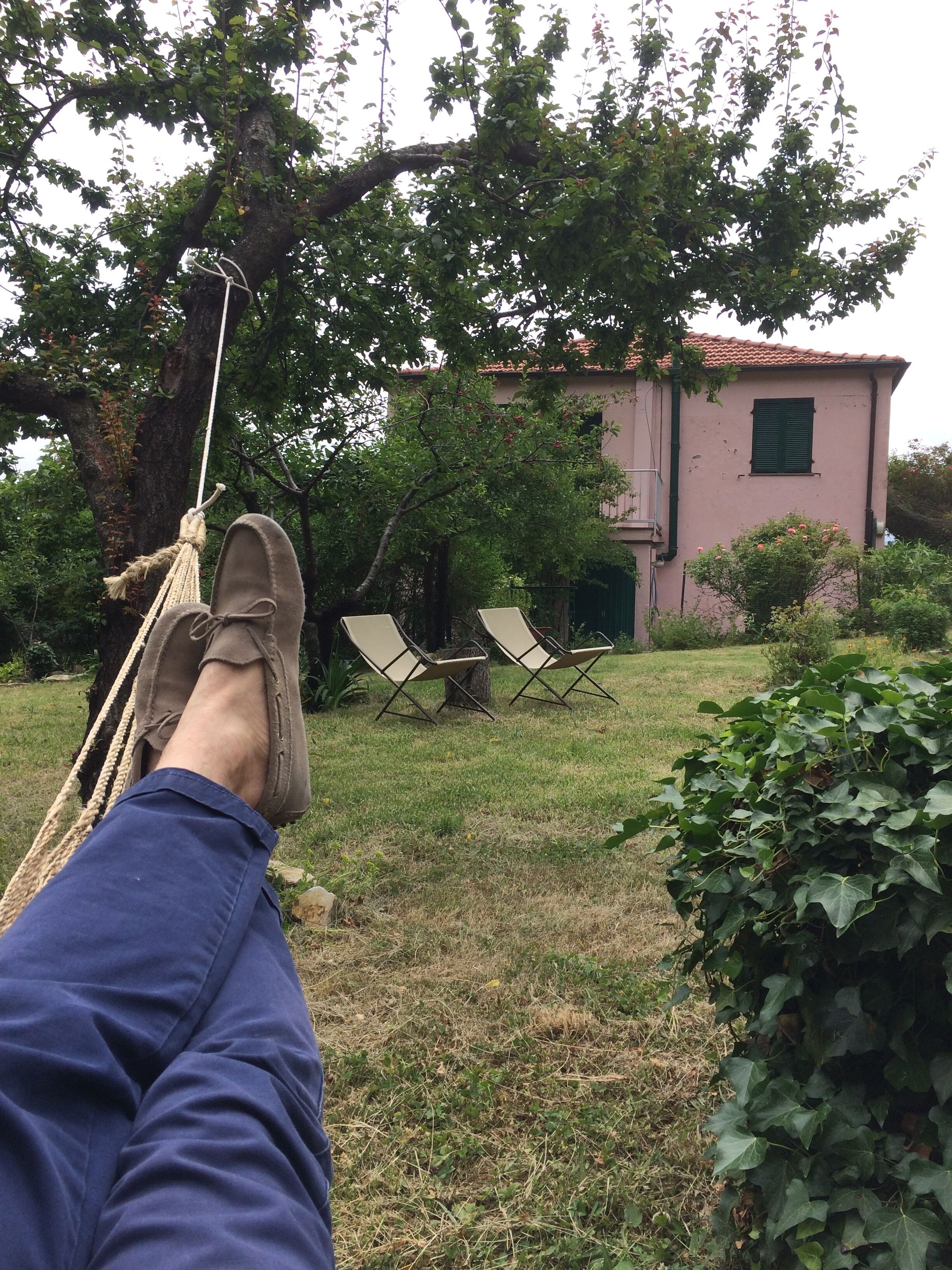 Una hamaca atada a un árbol, dos sillas de jardín y un edificio con fachada rosa al fondo.