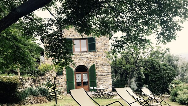 A stone building with green shutters, a lawn with lounge chairs, and a small table.