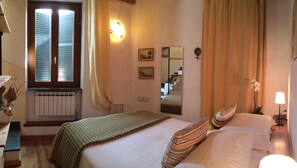 A hotel room with a bed, a window with blinds, a radiator, a mirror, a lamp, and a plant.