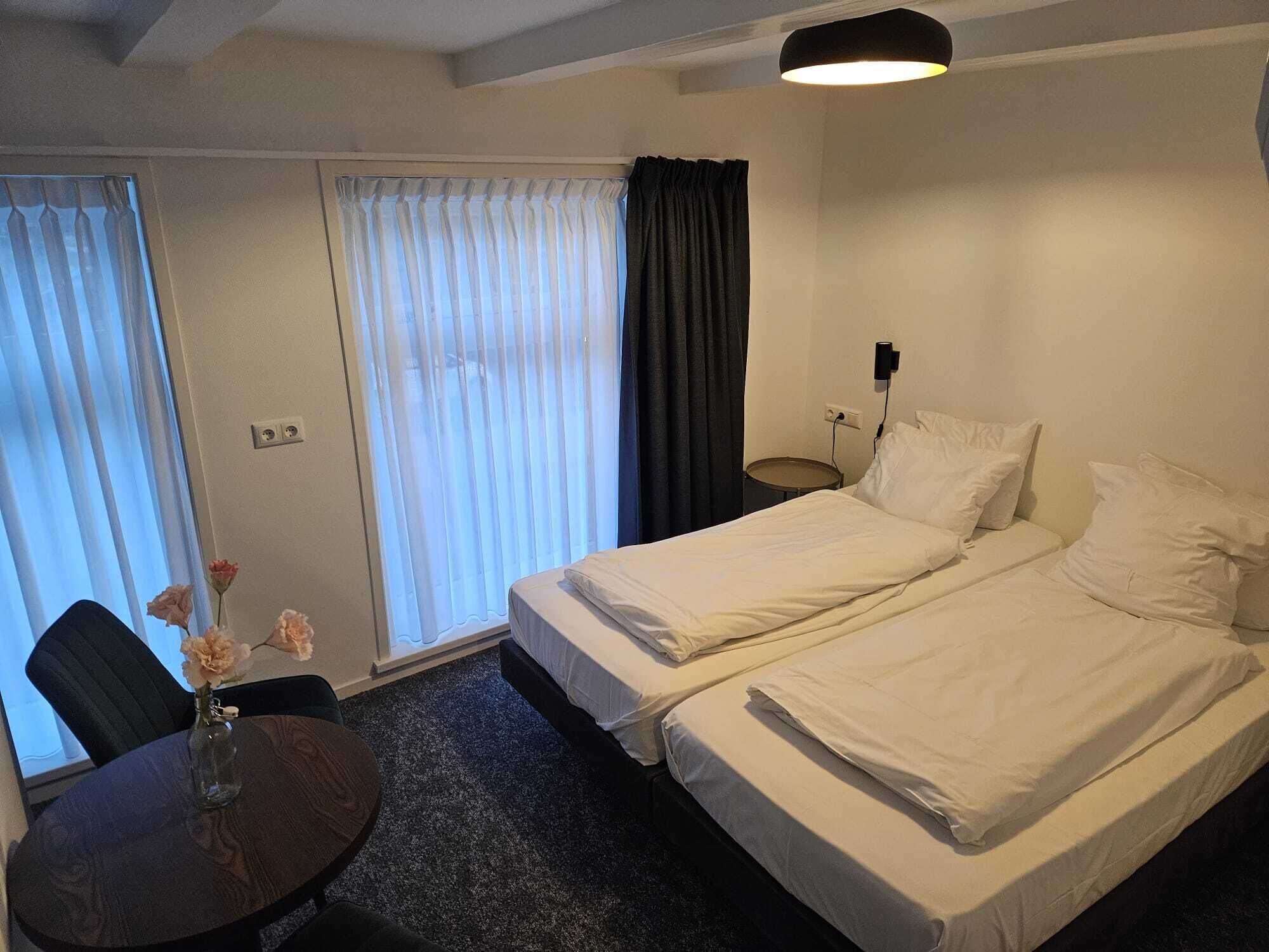 A hotel room with two beds, a small table with a vase of flowers, a chair, a round table, and a window with curtains.
