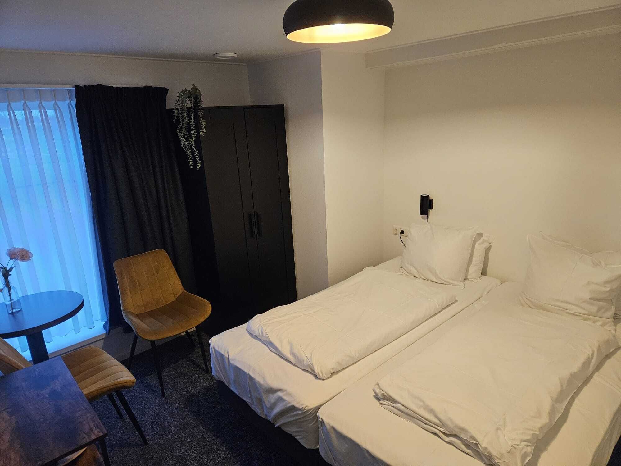 A hotel room with two single beds, a small table with a chair, a dark-colored wardrobe, and a window with curtains.
