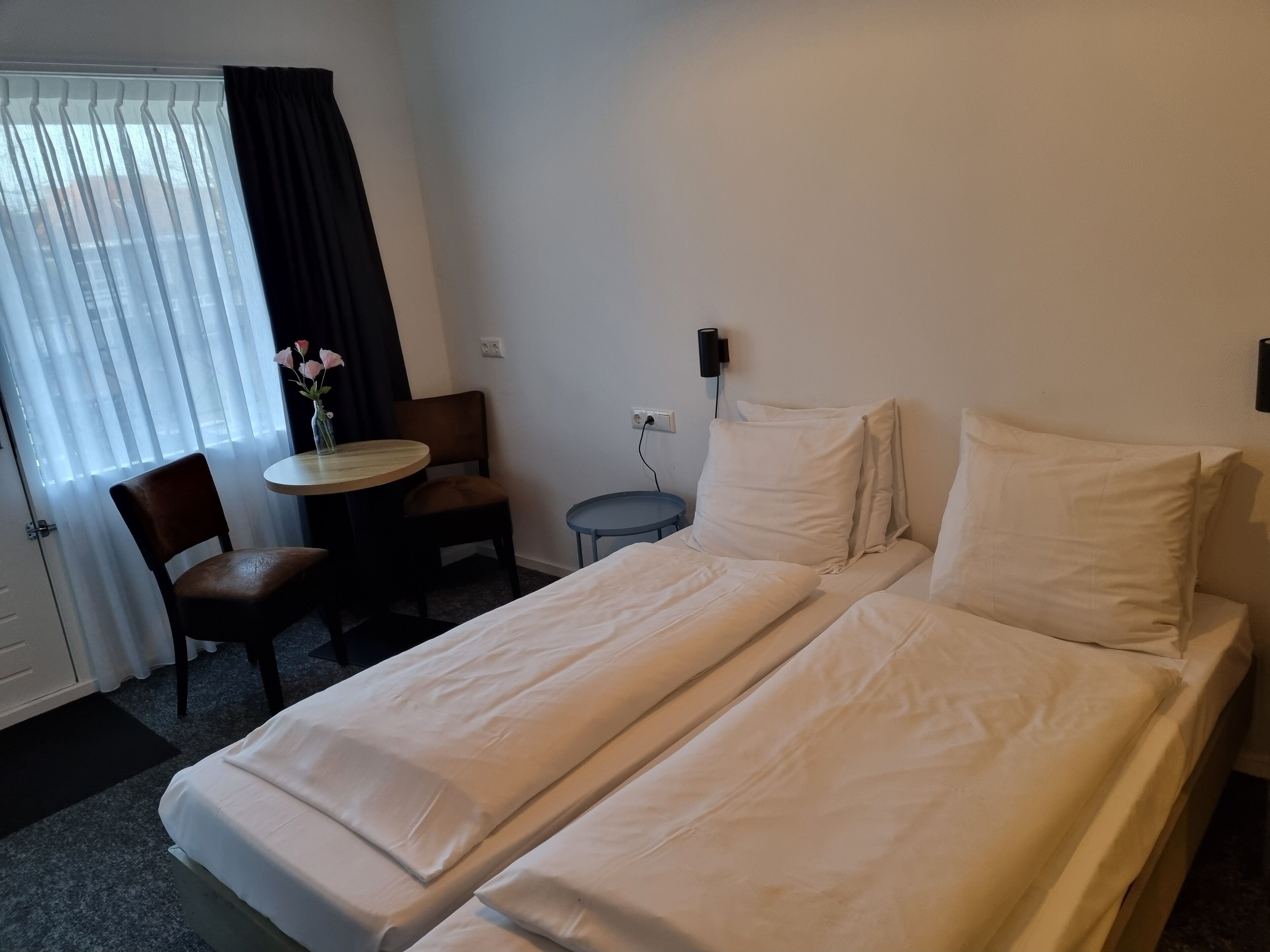 A hotel room with a double bed, a small round table with a chair, and a window with curtains.