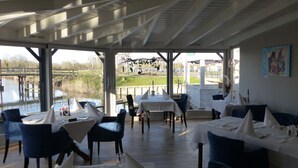 Un área de comedor al aire libre con mesas y sillas, vista a un río y un puente, y un cuadro en la pared.