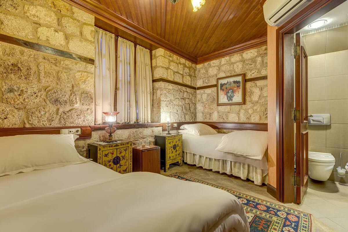 Una habitación con paredes de piedra, techo de madera, dos camas, una mesita de noche con lámpara, un cuadro y un baño con inodoro y lavamanos.