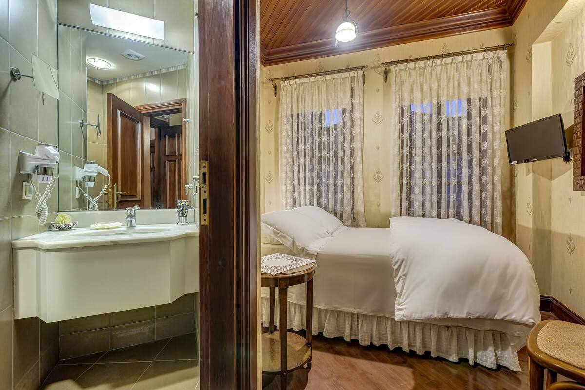 Habitación de hotel con una cama grande, una mesita de noche, un espejo, un lavamanos y un televisor montado en la pared.