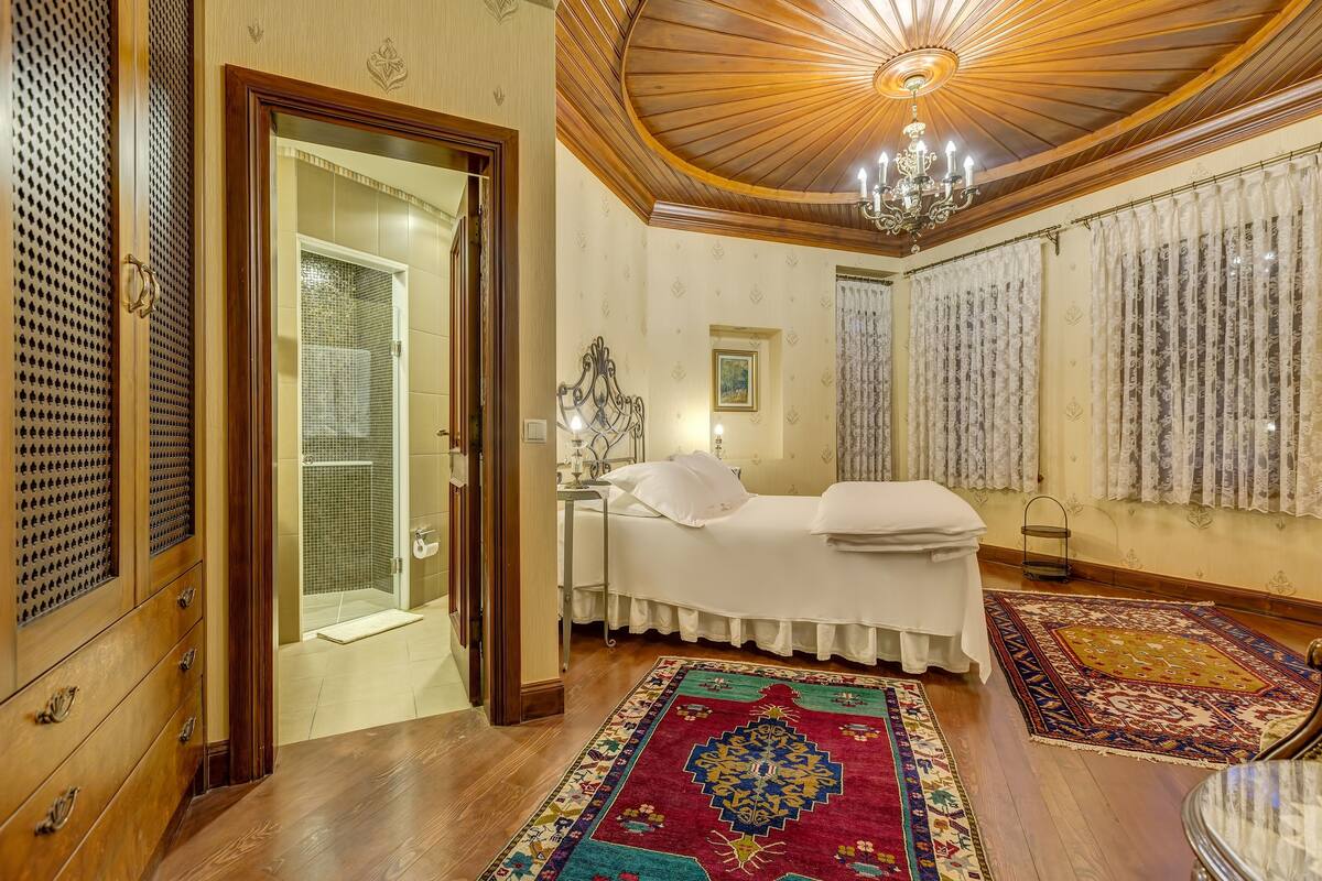 Un dormitorio tradicional con techo de madera, una cama, mesita de noche, lámpara de araña y cortinas estampadas.