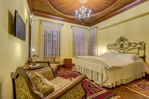 Un dormitorio con techo de madera, una araña, cama con ropa de cama blanca, dos sillones tapizados, un televisor y cortinas estampadas.