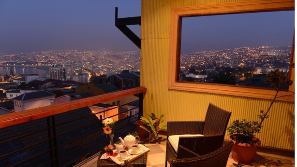 Un balcón con mobiliario de mimbre, una mesa con juego de té y vista a un paisaje urbano al anochecer.