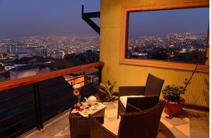 Un balcón con mobiliario de mimbre, una mesa con juego de té y vista a un paisaje urbano al anochecer.