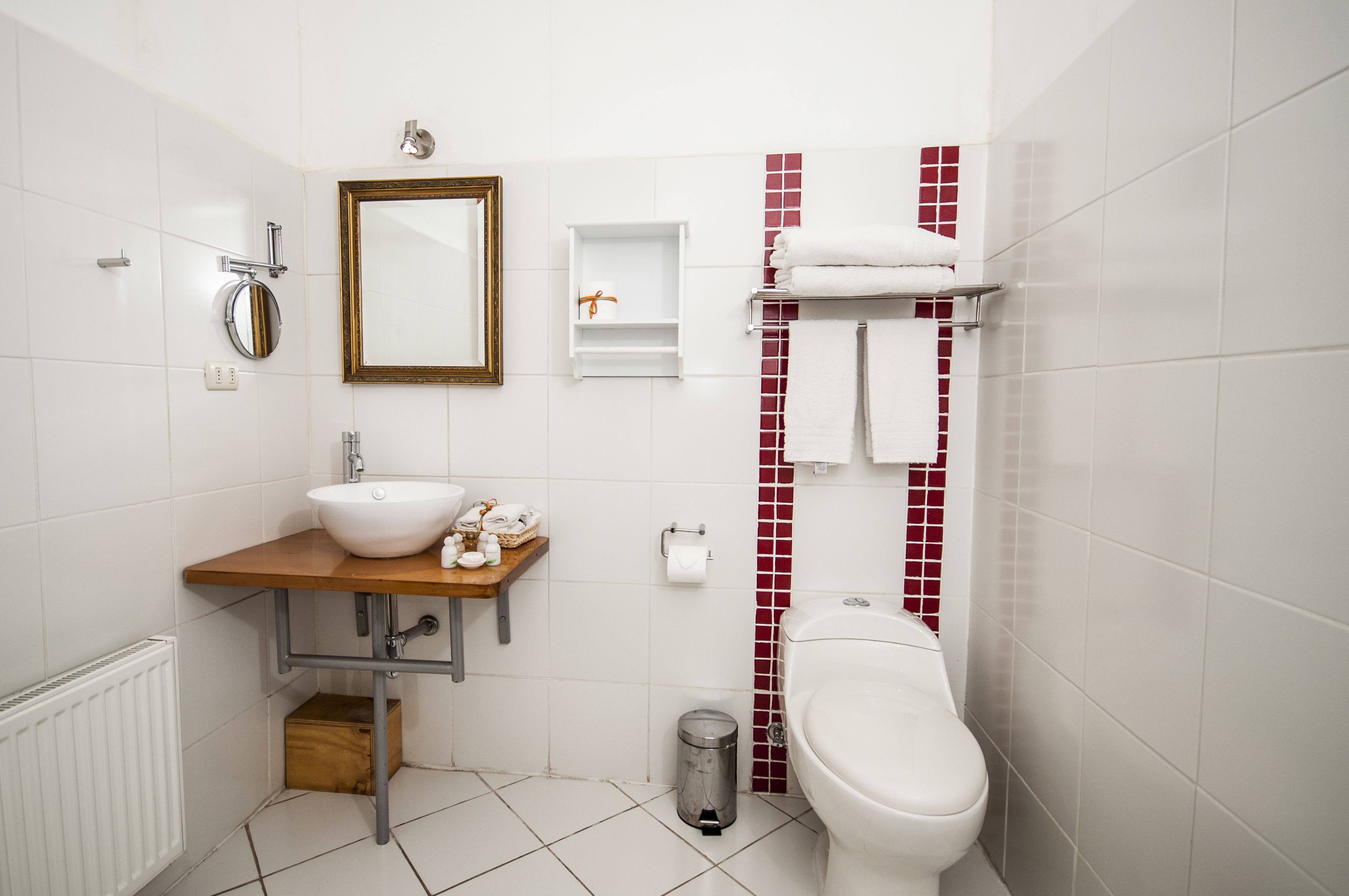 Une salle de bain moderne avec un lavabo blanc, un miroir et des toilettes. On y trouve une petite poubelle et une étagère avec des serviettes.