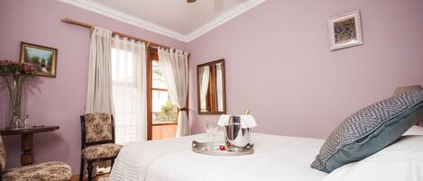 A bedroom with a bed, a chair, a bedside table with a vase, curtains, a ceiling fan, and a framed picture on the wall.