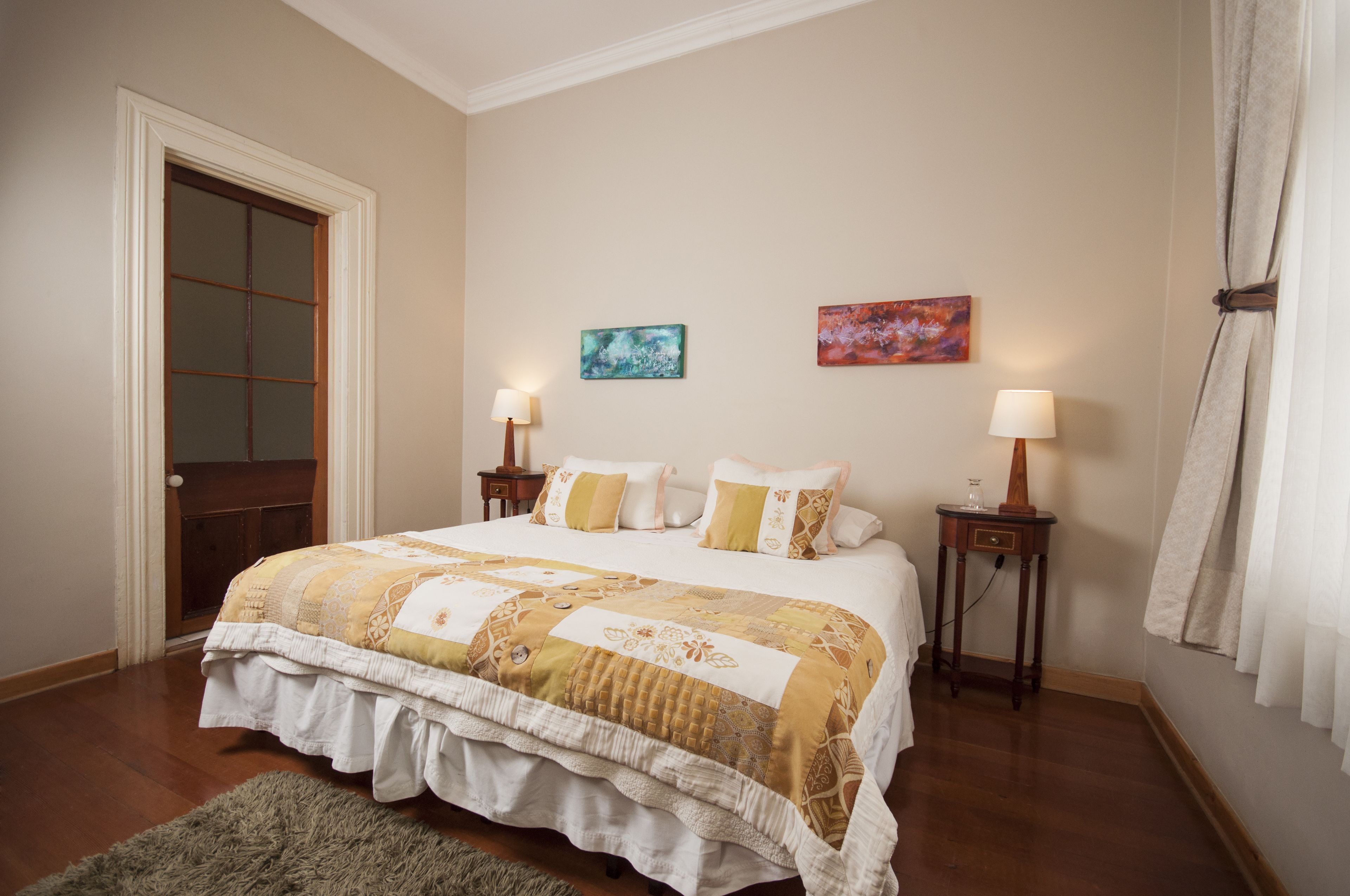 Un dormitorio con una cama grande, dos mesitas de noche con lámparas, una puerta y dos cuadros colgados en la pared.