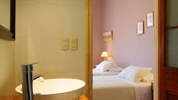 Un baño con un lavabo grande, un espejo y una encimera de madera. Se ve una cama a través de una puerta abierta en la habitación contigua.