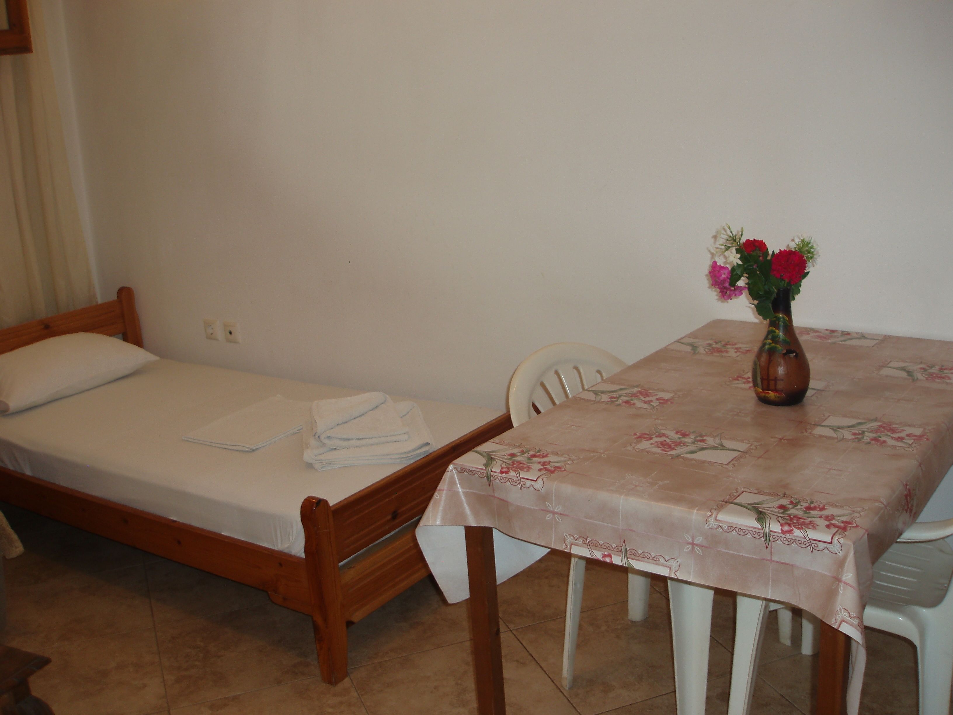 Una habitación pequeña con una cama individual, una mesa con mantel floreado y un florero con flores.