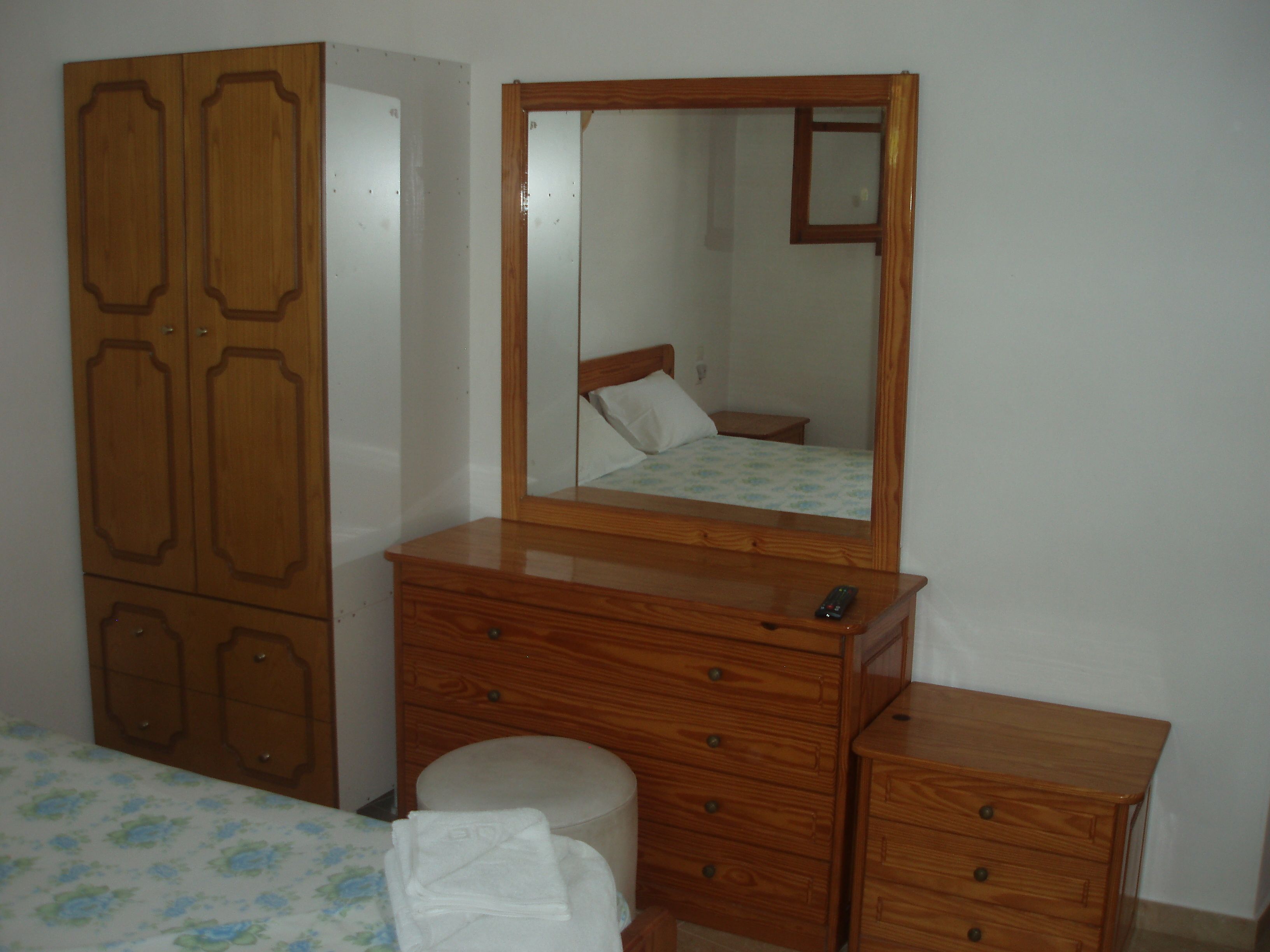Quarto com guarda-roupa de madeira, penteadeira, espelho, cama com colcha florida e mesa de cabeceira.