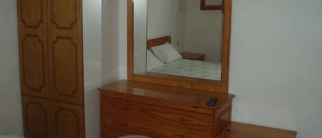 Quarto com guarda-roupa de madeira, penteadeira, espelho, cama com colcha florida e mesa de cabeceira.
