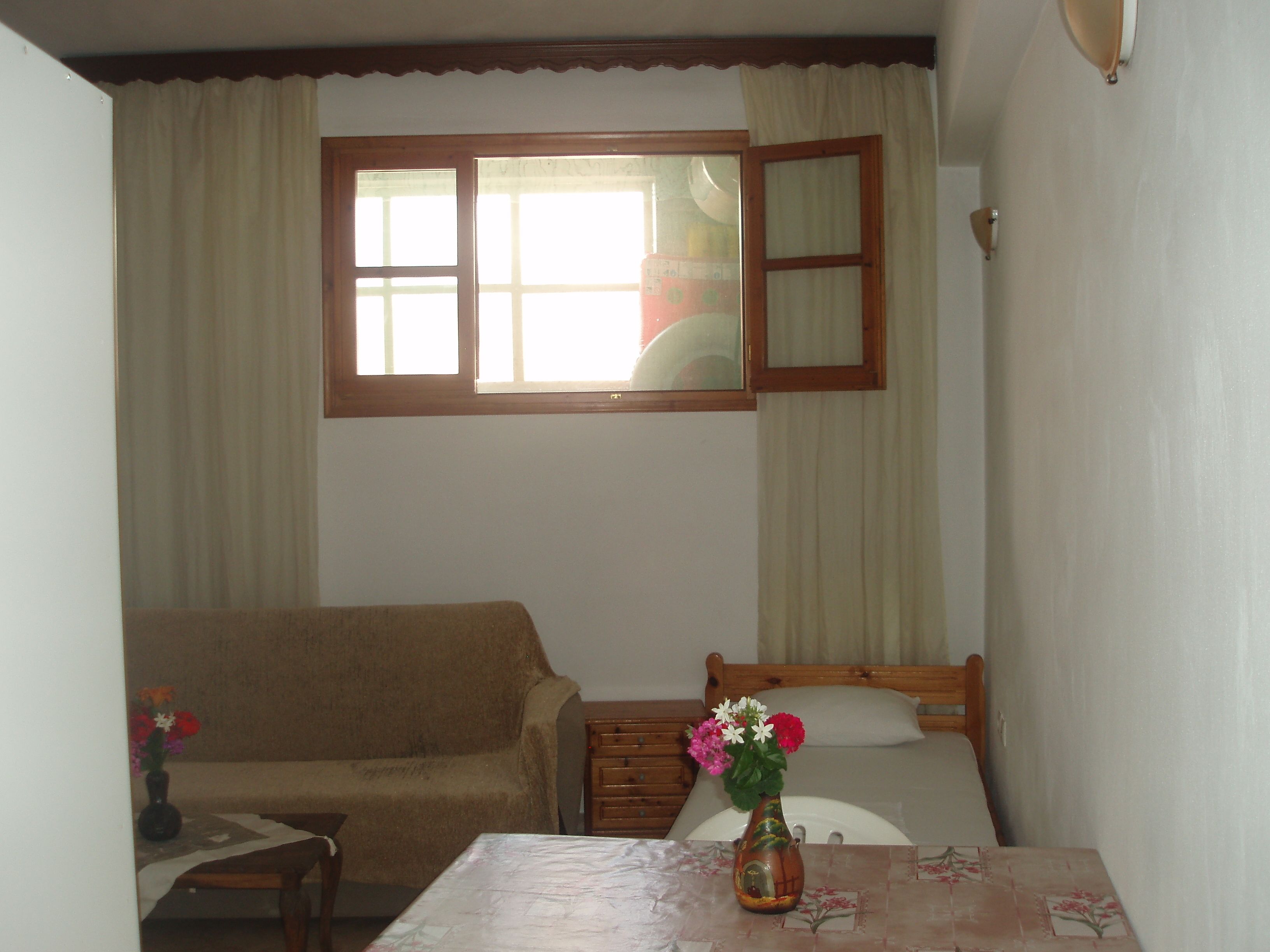 Ein kleines, gemütliches Zimmer mit einem Einzelbett, einem Holzstuhl, einem kleinen Tisch und einem Fenster mit Vorhängen.