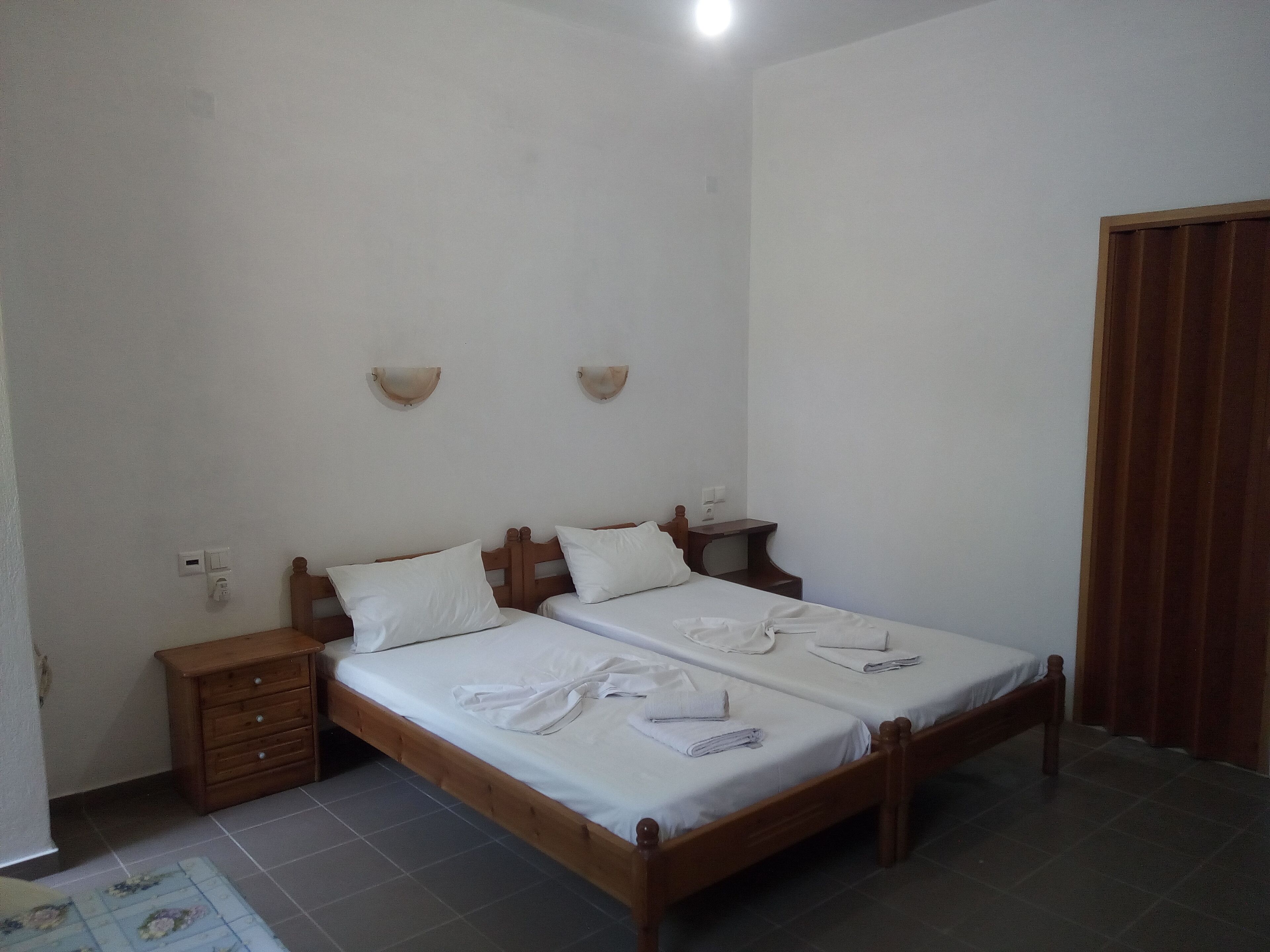 Una habitación sencilla y luminosa con cama doble, cabecera de madera, dos mesitas de noche y una cómoda pequeña.
