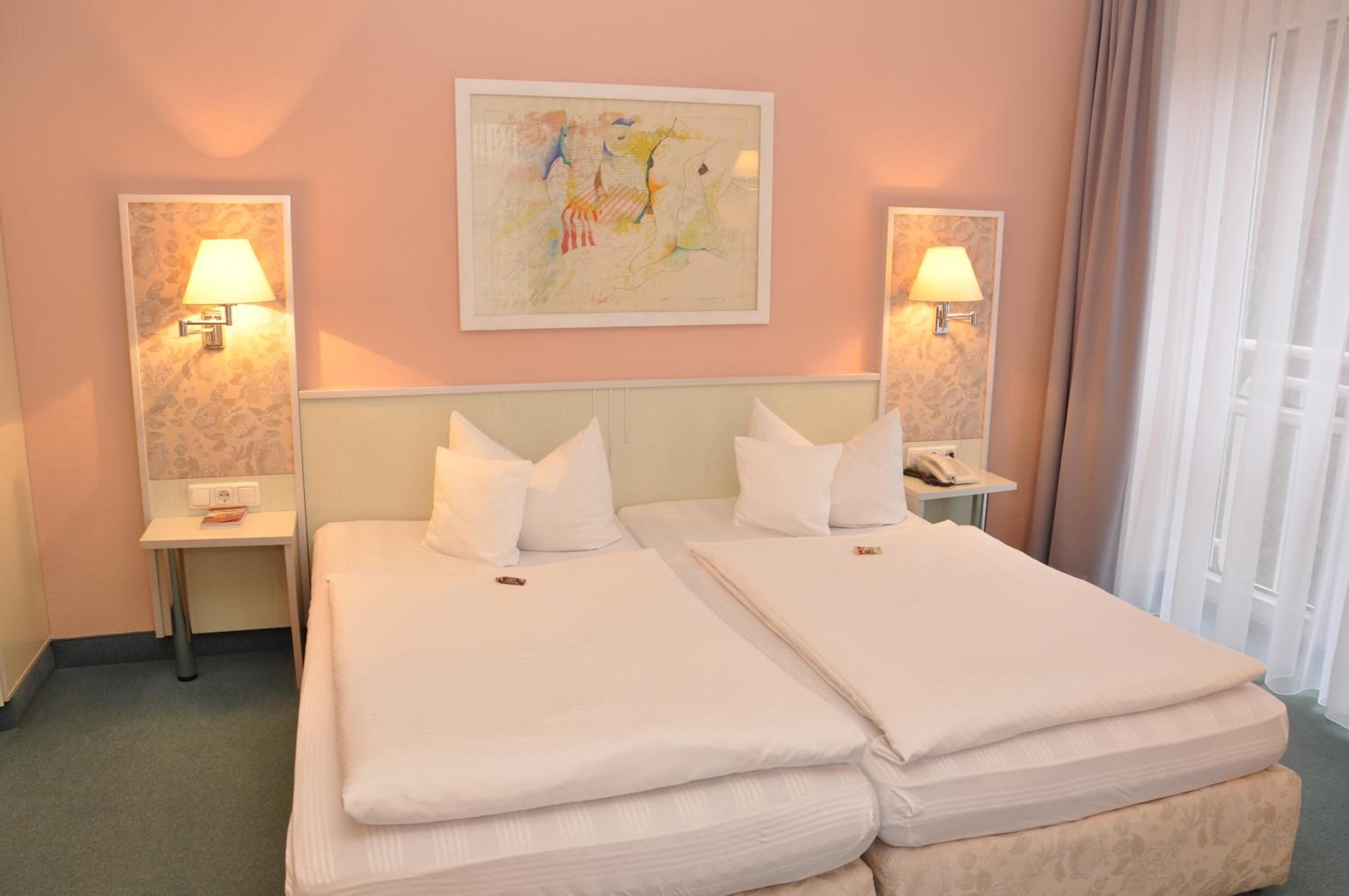 A hotel room with two beds, a nightstand with a lamp, a painting on the wall, and a window with curtains.