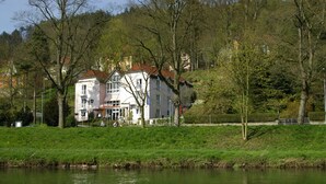 A hotel with a red roof and white walls, located beside a river with lush greenery and trees. - Altstadthotel An Der Werra (Meiningen)