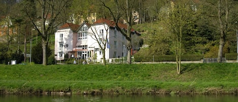 A hotel with a red roof and white walls, located beside a river with lush greenery and trees.