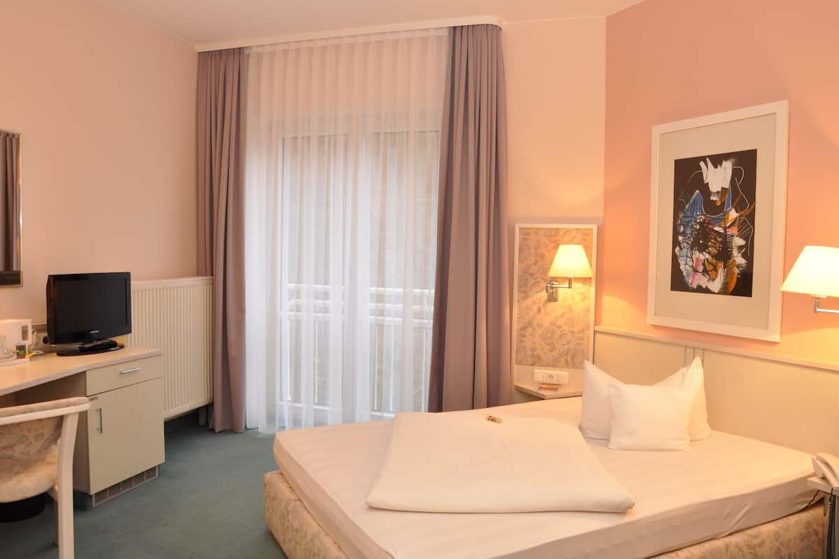 A hotel room with a bed, a desk with a computer, a lamp, and a painting on the wall.