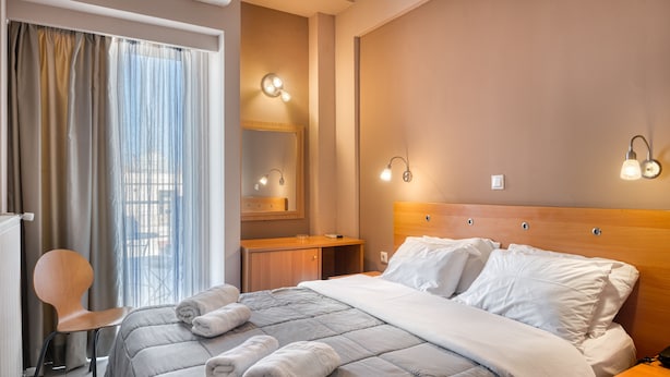 A hotel room with a bed, a wooden nightstand, a chair, a desk, and a mirror. - Diros Hotel (Athens)