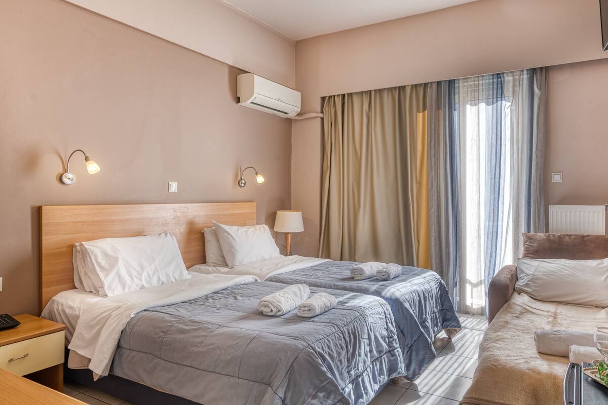 Quarto de hotel com duas camas, uma mesa de cabeceira com abajur, unidade de ar condicionado e janela com cortinas.