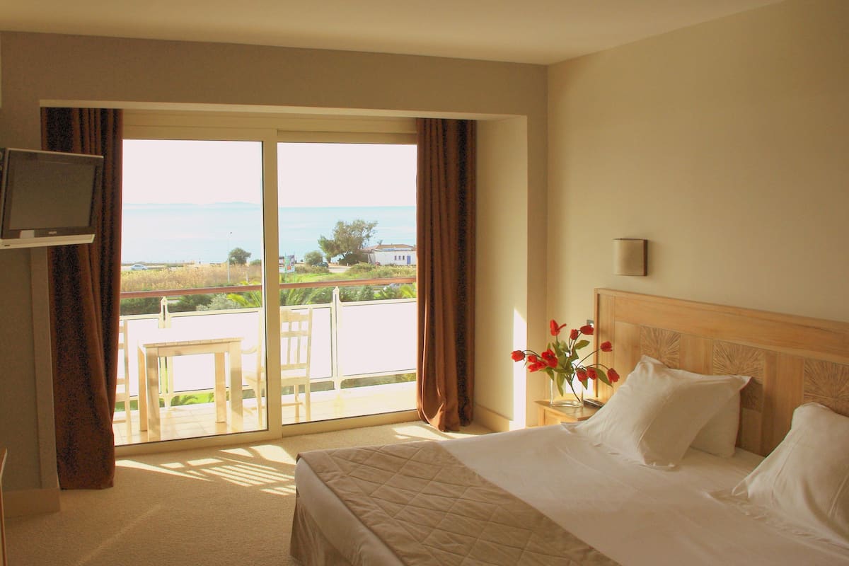Ein Hotelzimmer mit Bett, Fernseher, Nachttisch, Blumenvase und Blick ins Freie.