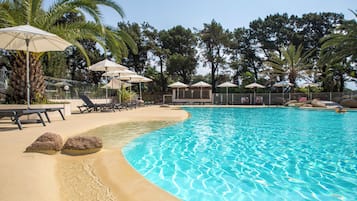 A swimming pool with lounge chairs and umbrellas, surrounded by palm trees and a wooden fence.