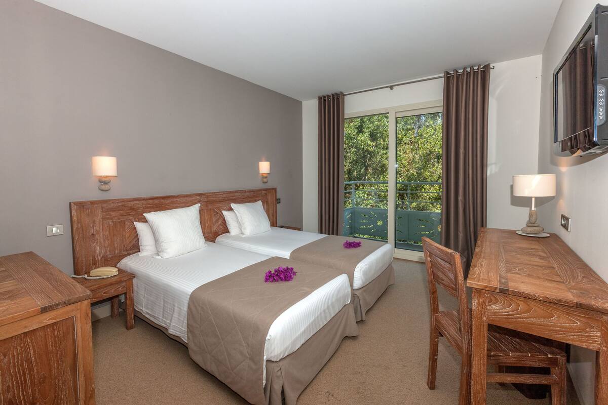 Ein Hotelzimmer mit zwei Betten, einem Holztisch, einem Fernseher und einem Balkon mit Vorhängen.