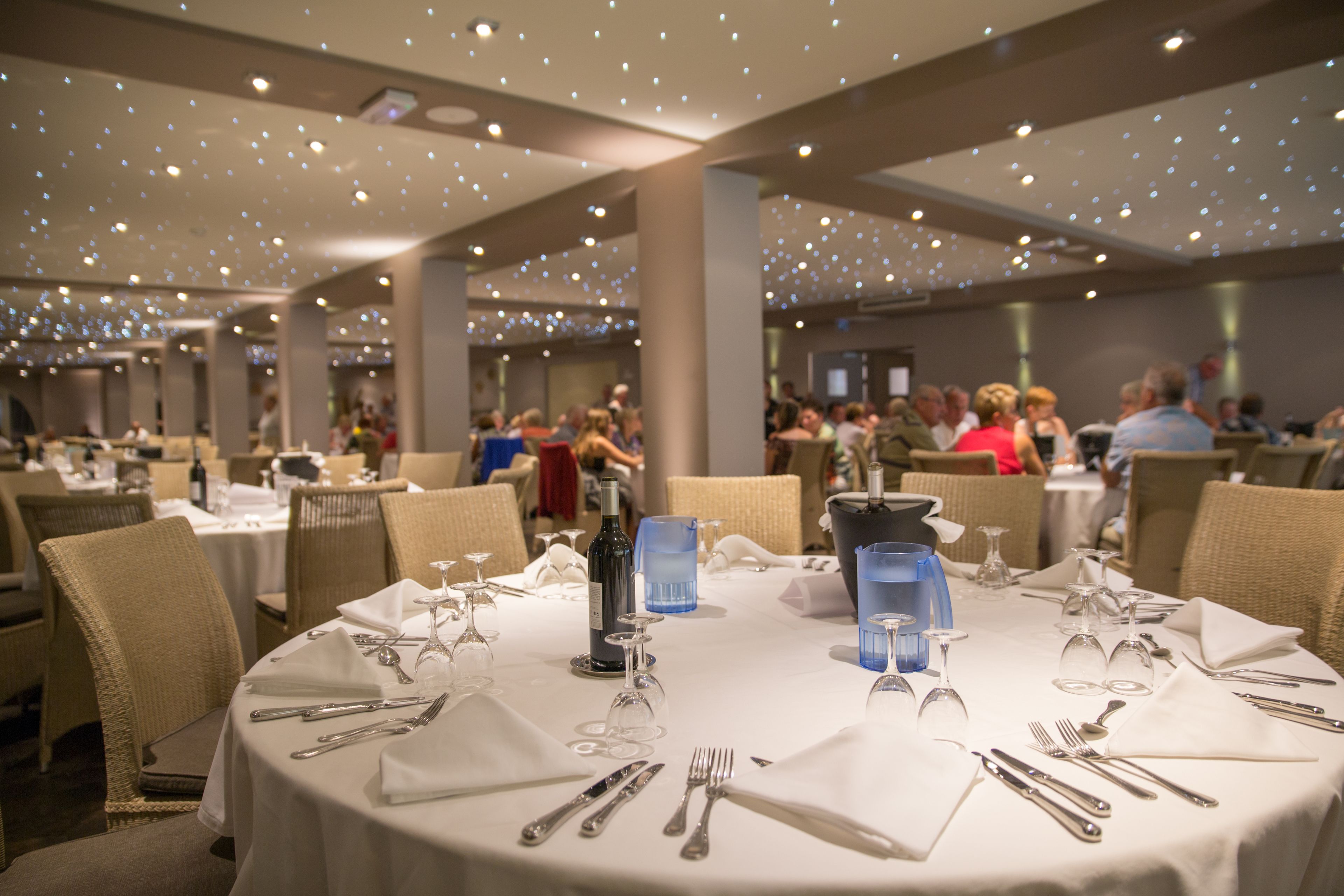 A dining area with round tables set for a meal, featuring white tablecloths, silverware, and blue water glasses.