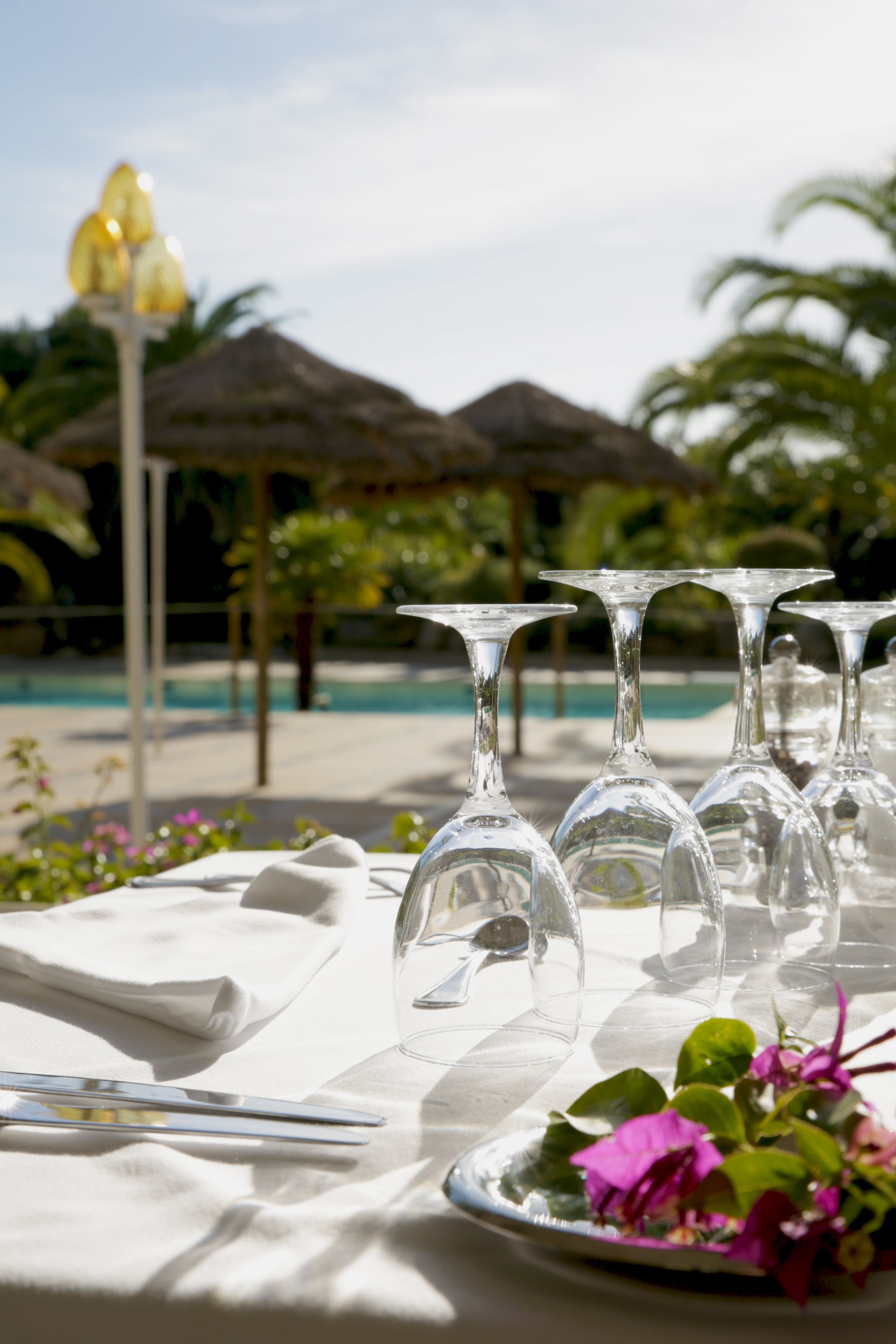 Mesa dispuesta con copas de vino, un plato con flores y una servilleta, con una piscina y sombrillas al fondo.