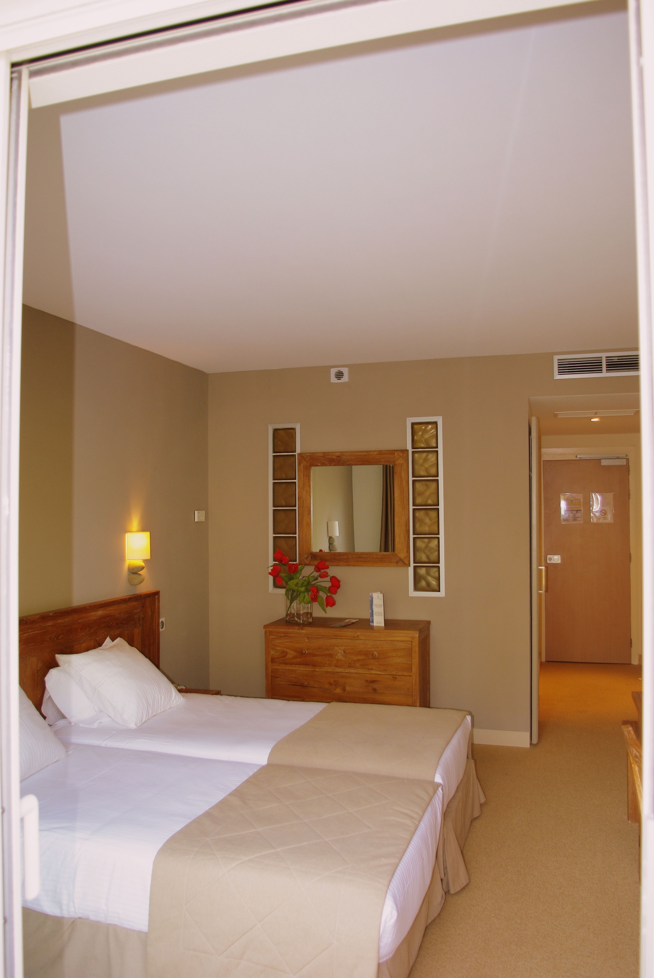 Une chambre d’hôtel avec deux lits, une commode en bois, un miroir et un vase avec des fleurs.
