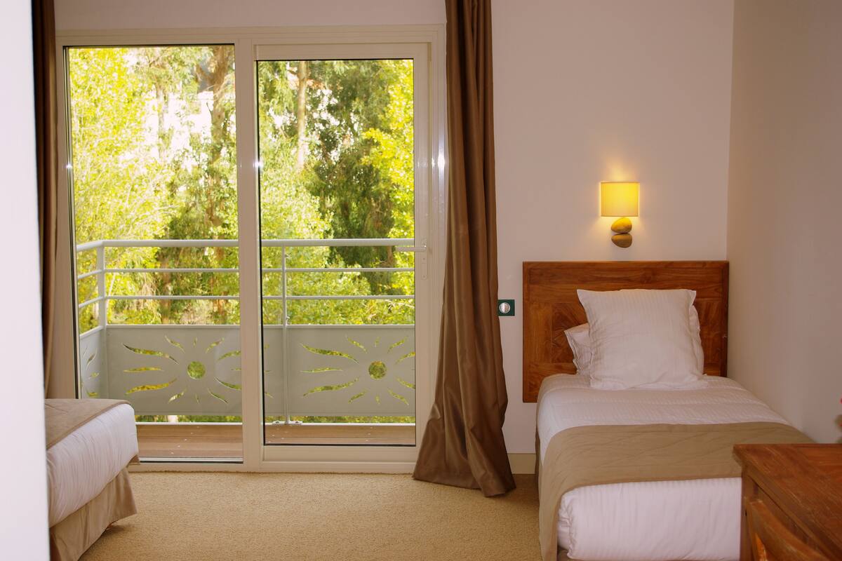 Ein Hotelzimmer mit Bett, Nachttisch, Lampe, Fenster mit Blick auf Grünflächen und einer Schiebetür zum Balkon.