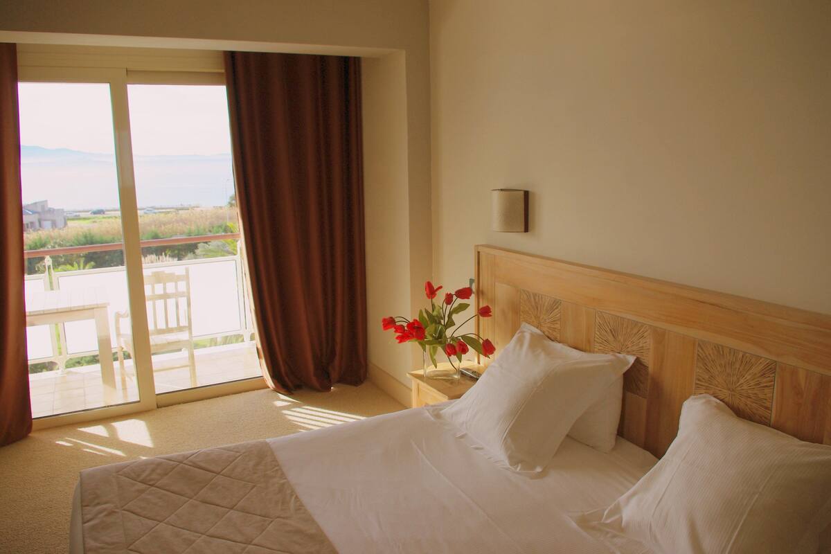 Ein Hotelzimmer mit Bett, Nachttisch mit Blumenvase, Lampe und Blick auf einen Balkon mit Tisch und Stühlen.