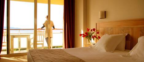 Quarto de hotel com uma cama, uma mesa de cabeceira com um vaso de flores vermelhas e vista para uma varanda com uma pessoa ao lado do corrimão.