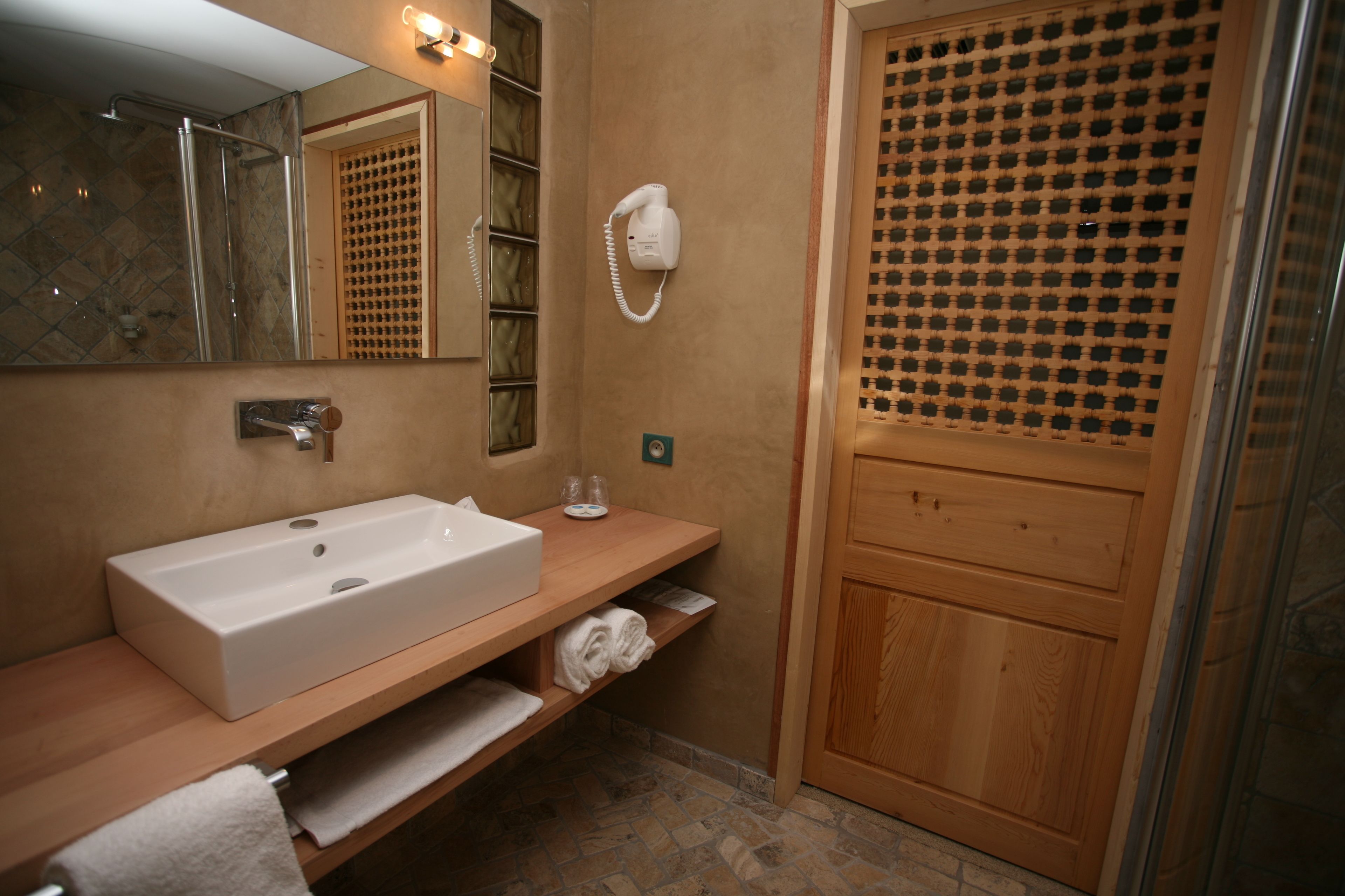 A bathroom with a large white sink, a mirror, a towel rack, a hairdryer, and a wooden door with a patterned glass panel.