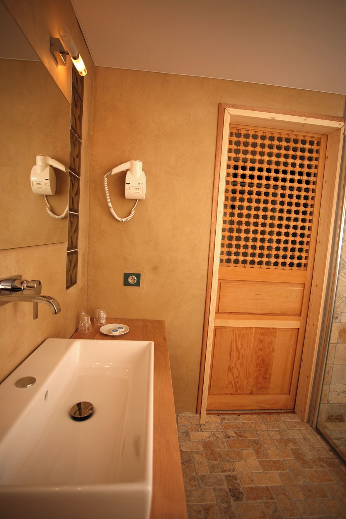 A bathroom with a large sink, two hair dryers mounted on the wall, a wooden door with a lattice design, and a tiled floor.
