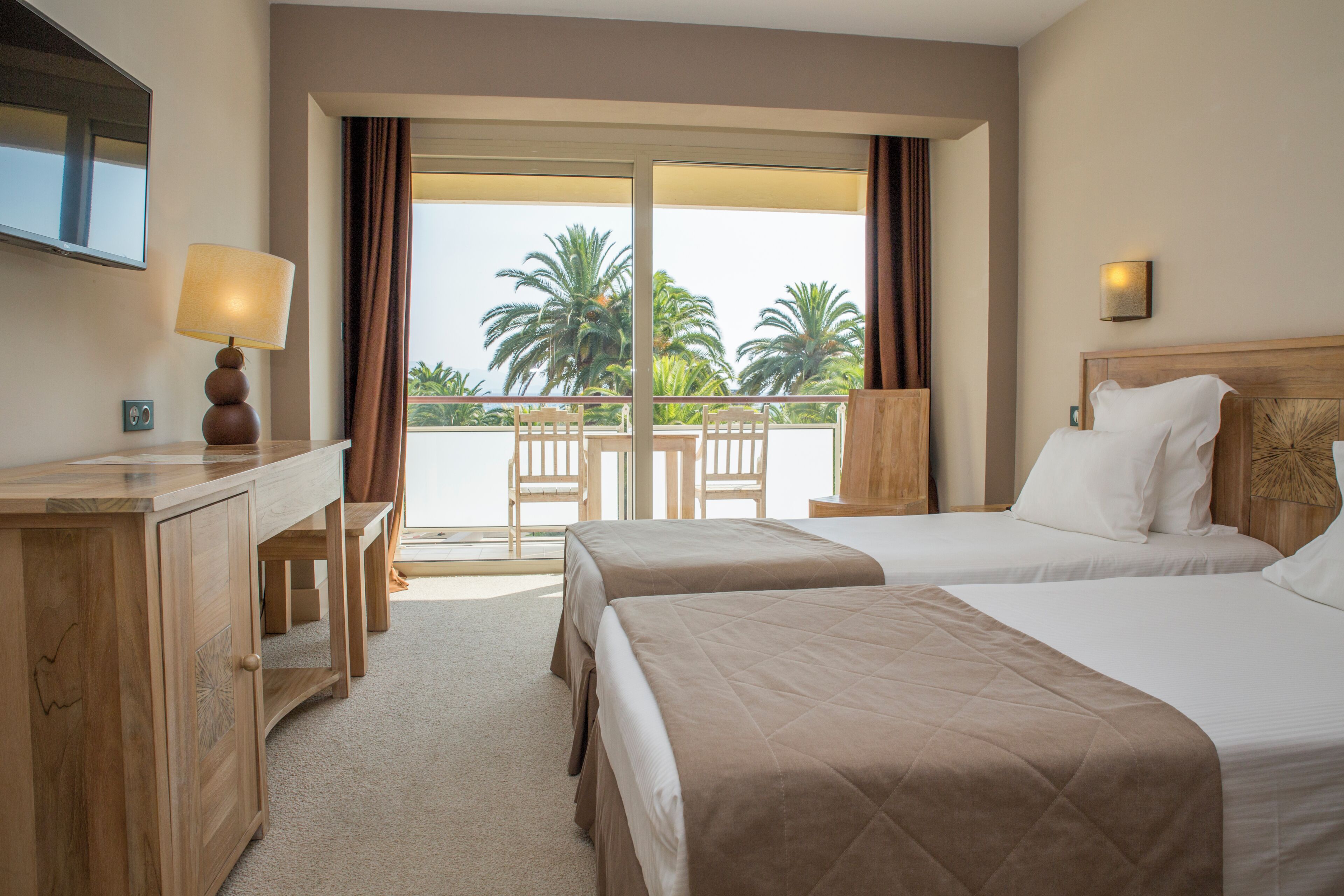 A hotel room with two beds, a desk, a chair, and a balcony with a view of palm trees.