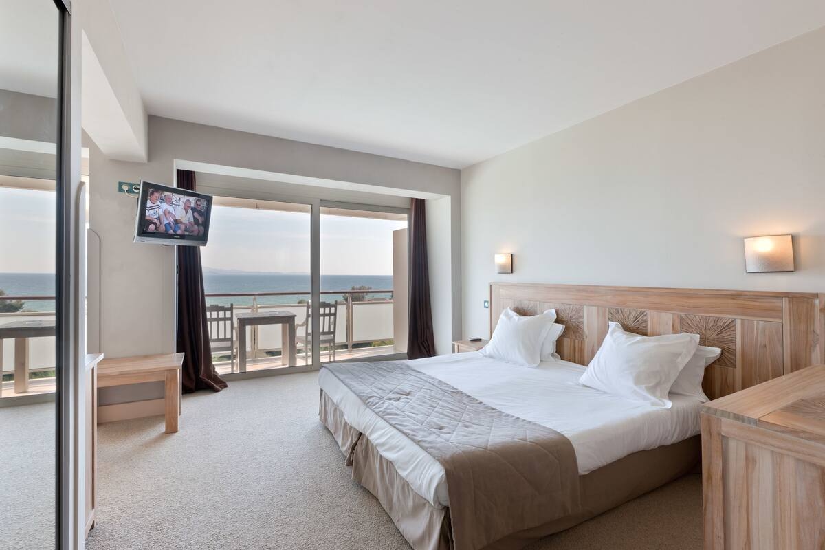 Ein Hotelzimmer mit einem großen Bett, einem an der Wand befestigten Fernseher, einem Balkon mit Tisch und Stühlen und Blick auf das Meer.