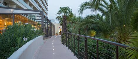 A pathway with a railing, surrounded by greenery and palm trees, leading to a building with multiple floors and large windows.