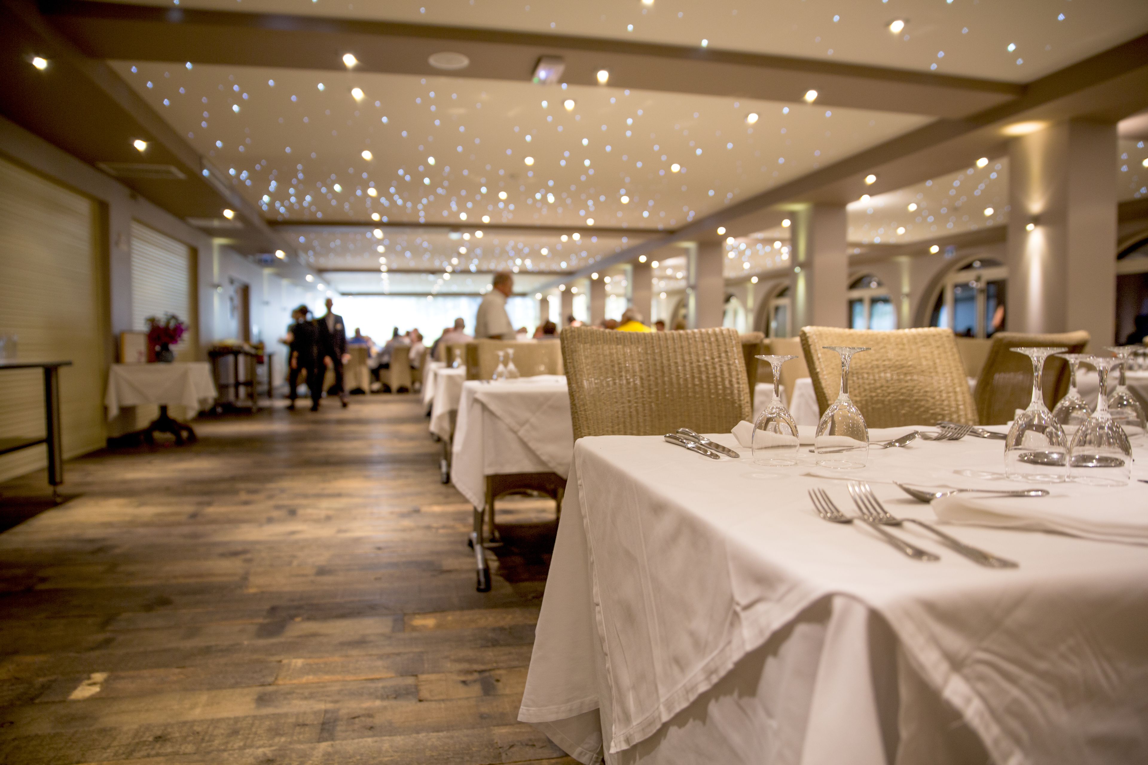 A long dining hall with tables set for a meal, featuring white tablecloths and wicker chairs.