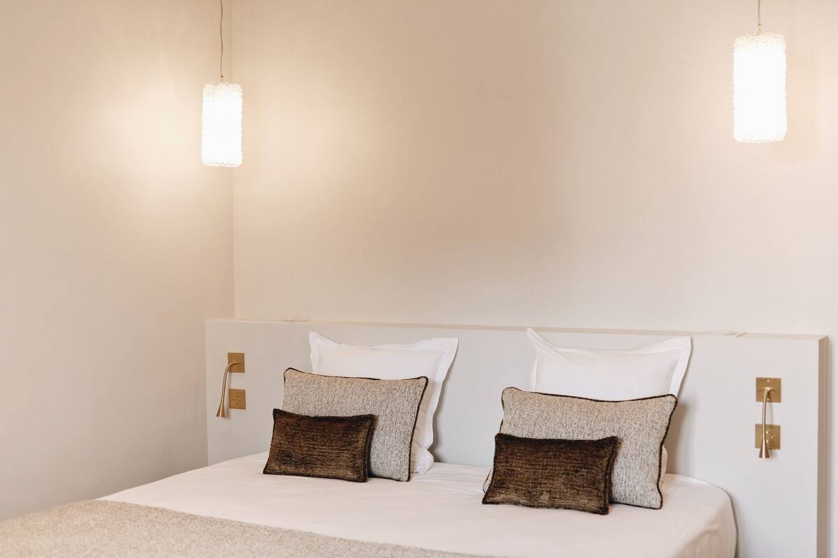Una cama bien hecha con dos almohadas, una mesita de noche y dos lámparas fijadas a la pared.
