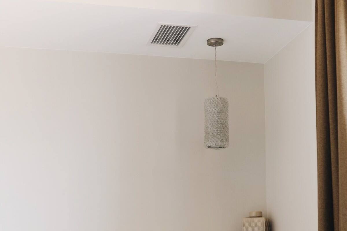 Un dormitorio con cama, almohadas, una lámpara colgante, una luz instalada en la pared y un respiradero en el techo.
