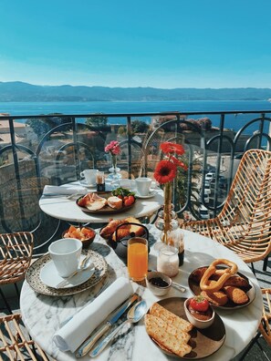 A balcony table with breakfast items, a wicker chair, and a scenic view of the sea and mountains. - Hotel San Carlu Citadelle (Ajaccio)