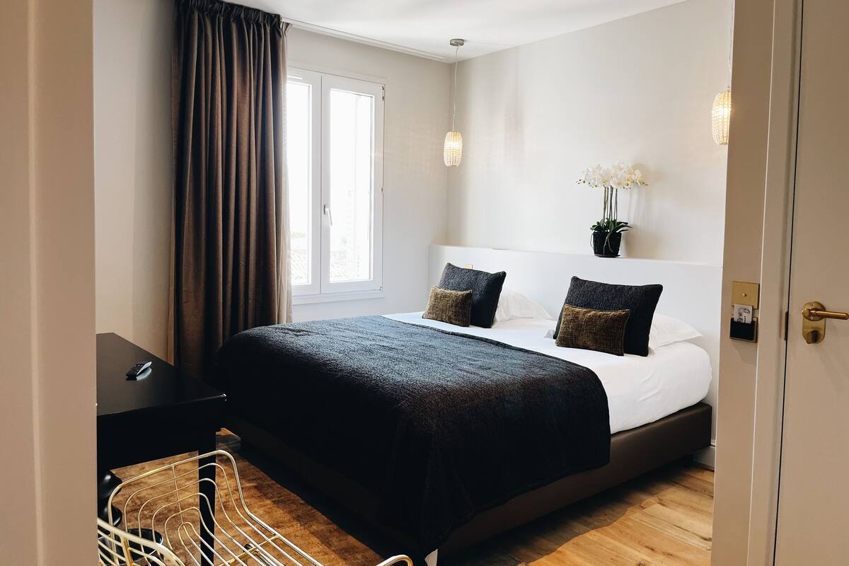Habitación de hotel con una cama grande, una mesita de noche, una silla, un ventanal con cortinas y una planta en maceta sobre la mesita.