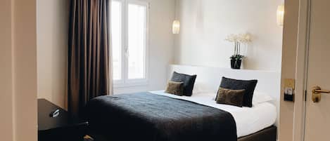A hotel room with a large bed, a bedside table, a chair, a window with curtains, and a potted plant on the nightstand.