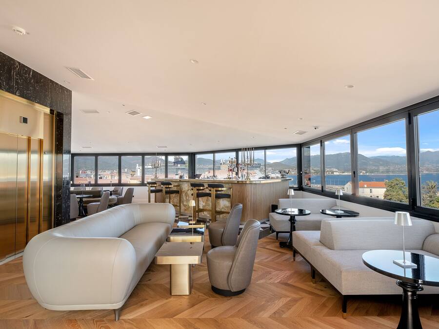 A modern hotel lobby with a high ceiling, large windows, a bar area, and a seating area with sofas and tables.