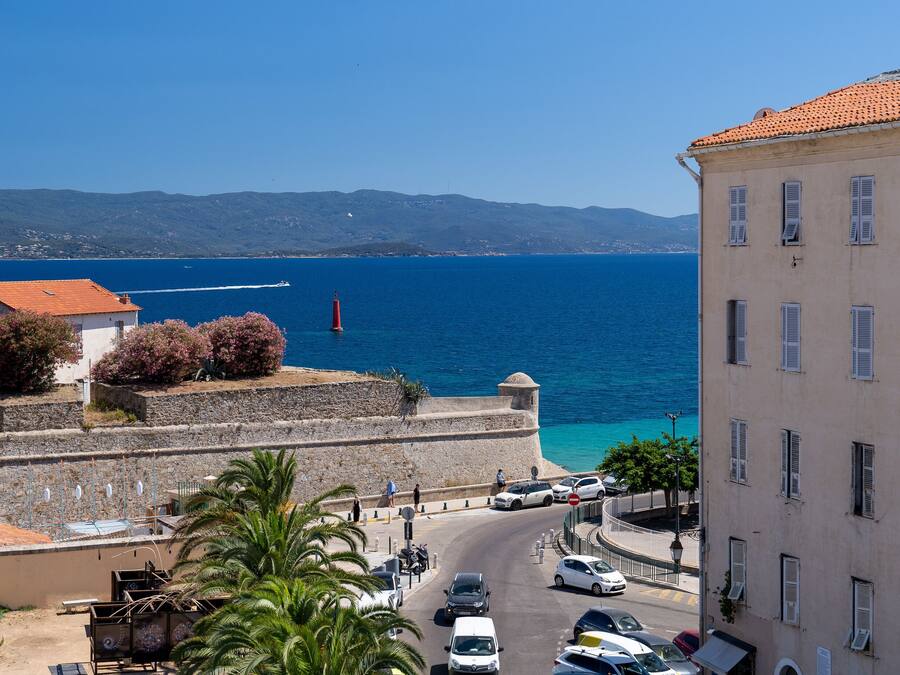 A coastal street with buildings, a clear blue sea, and a distant mountain range.